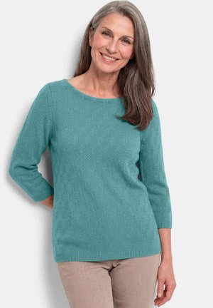 Lachende vrouw met lang grijs haar, gekleed in een teal gebreide trui met driekwart mouwen en beige broek tegen een effen achtergrond.