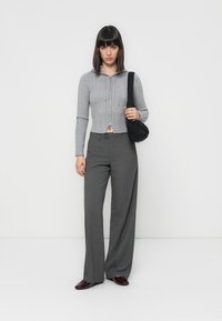 Donna con capelli scuri e lisci, che indossa un maglione grigio con zip, pantaloni larghi grigi, scarpe bordeaux e tiene in mano una borsa a tracolla nera.