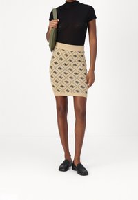 Femme portant un haut noir à manches courtes, une mini-jupe beige avec un motif en losanges noirs, des mocassins noirs et portant un sac à épaule vert olive.