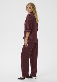Conjunto de satén burdeos de dos piezas que incluye una camisa de manga larga y pantalones sueltos. Corte ajustado con cuello y mangas con puños, complementado con tacones negros.
