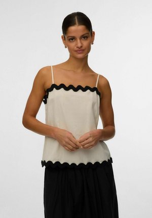 Femme aux cheveux foncés attachés, portant un haut beige sans manches avec une bordure noire ondulée et une jupe noire, debout devant un fond blanc uni.