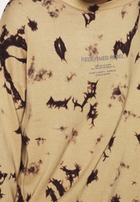 Sudadera de algodón beige con un patrón de teñido anudado negro. El texto dice "REDEFINED REBEL." Cuello redondo acanalado y tela texturizada.