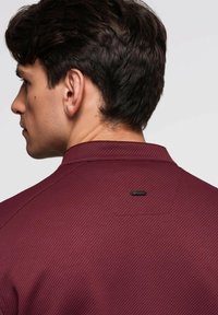 Burgunderfarbendes Poloshirt mit strukturiertem Muster, ausgestattet mit einem Kragen und einem kleinen schwarzen Logo-Tag auf der Rückseite, auf einem neutralen Hintergrund präsentiert.