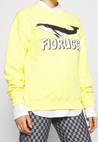 Sudadera amarilla con gráfico de ballena negra y texto "FIORUCCO". Hecha de una tela suave, cuenta con cuello redondo y puños acanalados.