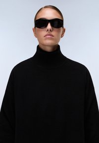 Musta ylipitkä pooloneule, jossa on ribattu kaulus, yhdistettynä paksuihin mustiin suorakulmaisiin aurinkolaseihin. Näkyvissä sileä kangasrakennetta.