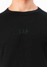 Gros plan sur une personne portant une chemise à manches longues noires texturée avec un petit logo GAP en relief sur la poitrine.
