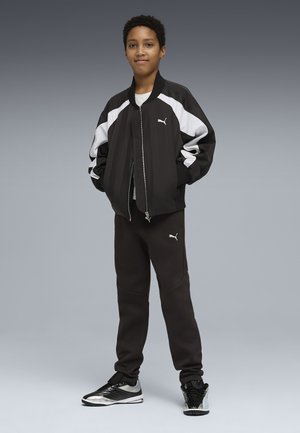 Barn står iført sort og hvid Puma træningsdragt med hænderne i lommerne og sorte og sølvfarvede sneakers, grå baggrund.