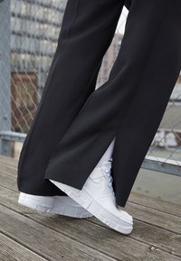 Sneaker bianchi con tomaia testurizzata e suola piatta, abbinati a pantaloni neri a gamba larga con spacco laterale. Superficie in legno sullo sfondo.