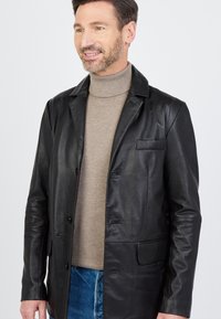 Czarna skórzana kurtka z ząbkowanym kołnierzem, przednimi kieszeniami i zapięciem na guziki. Noszona na beżowym swetrze z golfem i niebieskich dżinsach.