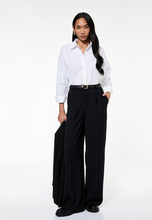 Kobieta nosząca białą koszulę na guziki włożoną w czarne szerokie spodnie z wysokim stanem, trzymająca czarną kurtkę w jednej ręce.