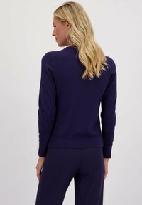 Paarse gebreide trui met een geribde zoom en manchetten, voorzien van knoopaccenten. De structuur lijkt zacht en glad.