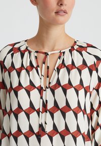 Vloeiende blouse met een geometrisch patroon in rood, zwart en wit. Sleutelgatnek met strikjes, lichtgewicht stof en geplooide halslijn detail.