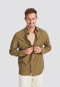 Khaki button-up shirt, long sleeves, soft fabric, relaxed fit, worn over a beige shirt, features a classic collar and brown buttons.