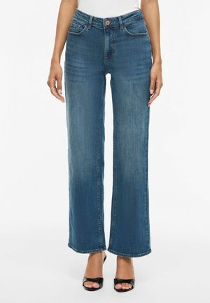 Ženska v modrih kavbojkah z visokim pasom in širokimi nogami ter črnih odprtih sandalih s peto stoji na belem ozadju.