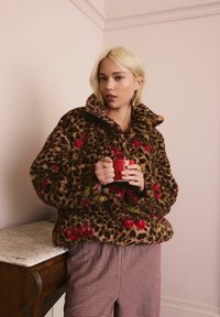 Polarowy pulower w print w panterkę z haftem kwiatowym, wysoki kołnierz, długie rękawy; model trzyma czerwony kubek, nosząc w kratkę spodnie.