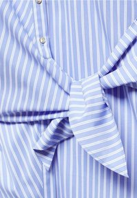 Chemise à rayures verticales bleu clair et blanches avec boutons et un détail en tissu noué à l'avant.