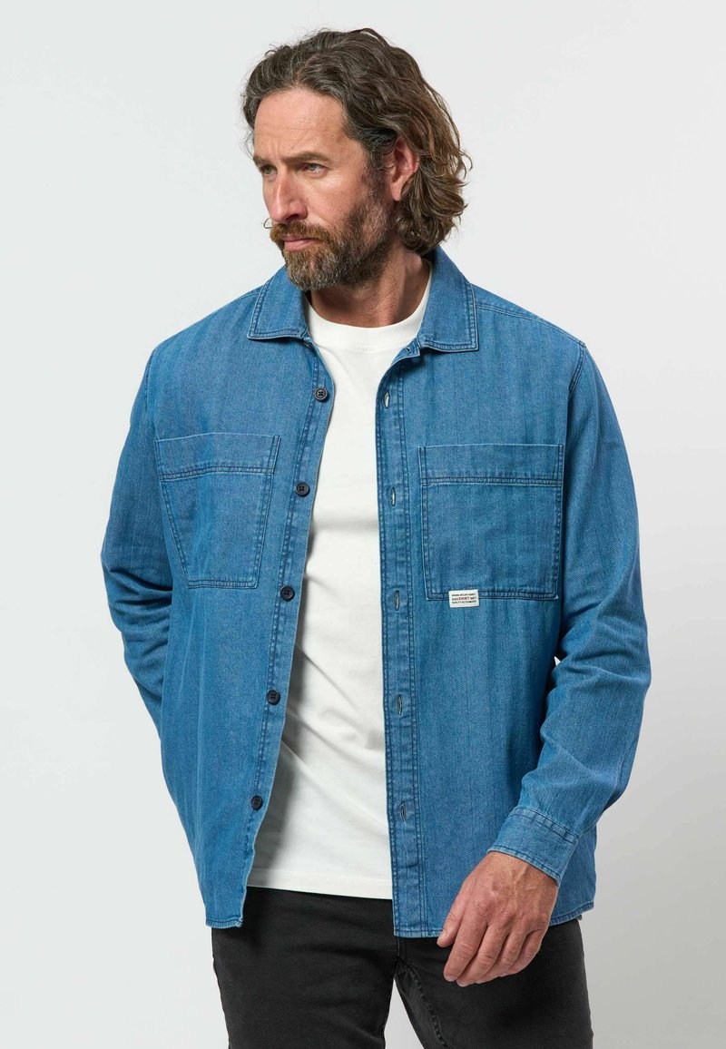 Homme aux cheveux bouclés et à la barbe, portant une chemise en jean bleue sur un t-shirt blanc, debout les mains dans les poches, regardant vers la gauche.