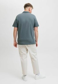 Polo gris à manches courtes, en tissu texturé, associé à un pantalon beige clair et des baskets blanches. Vue de dos, tourné à l'écart de l'objectif.