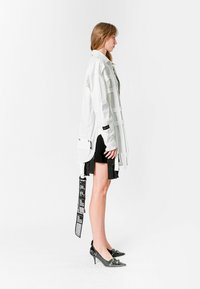 Chaqueta blanca de manga larga con múltiples bolsillos y correas, combinada con una falda negra con pliegues y tacones negros puntiagudos, sobre un fondo blanco.