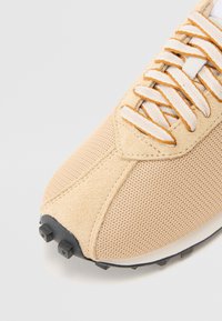 Beige träningssko med övre av mesh och mocka, vita och orange snörning, samt en svart gummisula med räfflade dobbar.