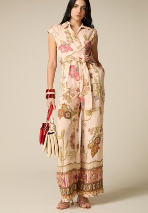 Mujer con mono floral beige de pierna ancha, sosteniendo bolsos rojos y beige, complementado con pulseras rojas y sandalias de tacón.