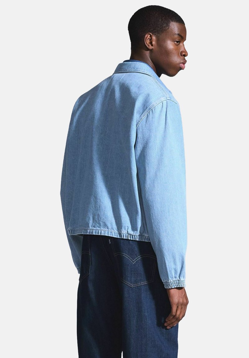 Hellblaues Denim-Jackett mit lässigem Schnitt, sichtbaren Nähten und elastischen Bündchen, getragen mit dunkelblauen Jeans, Ansicht von hinten.