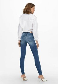 Bijela bluzica s složenim čipkastim detaljima, u kombinaciji s plavim skinny trapericama visokog struka. Model nosi bijele sandale s otvorenim prstima.