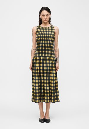 Kobieta z tatuażami w żółto-czarnej sukience midi w kratę bez rękawów i czarnych balerinach, stojąca na tle jednolitej białej ściany.