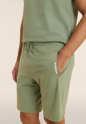 Uomo che indossa pantaloncini verde oliva con vita con coulisse e logo "FREDDY" sulla tasca, mano appoggiata all'interno della tasca.
