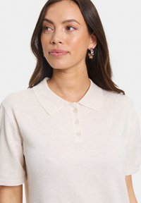 Beige polo-shirt med klassisk krage, fem knappar och korta ärmar. Tillverkad av ett texturerat tyg med ett subtilt mönster.