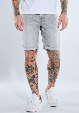 Mann trägt hellgraue, hochgekrempelte Jeansshorts, weiße Sneakers und hat sichtbare Bein-Tattoos mit Sonnen-, Schmetterlings- und Blumenmotiven.