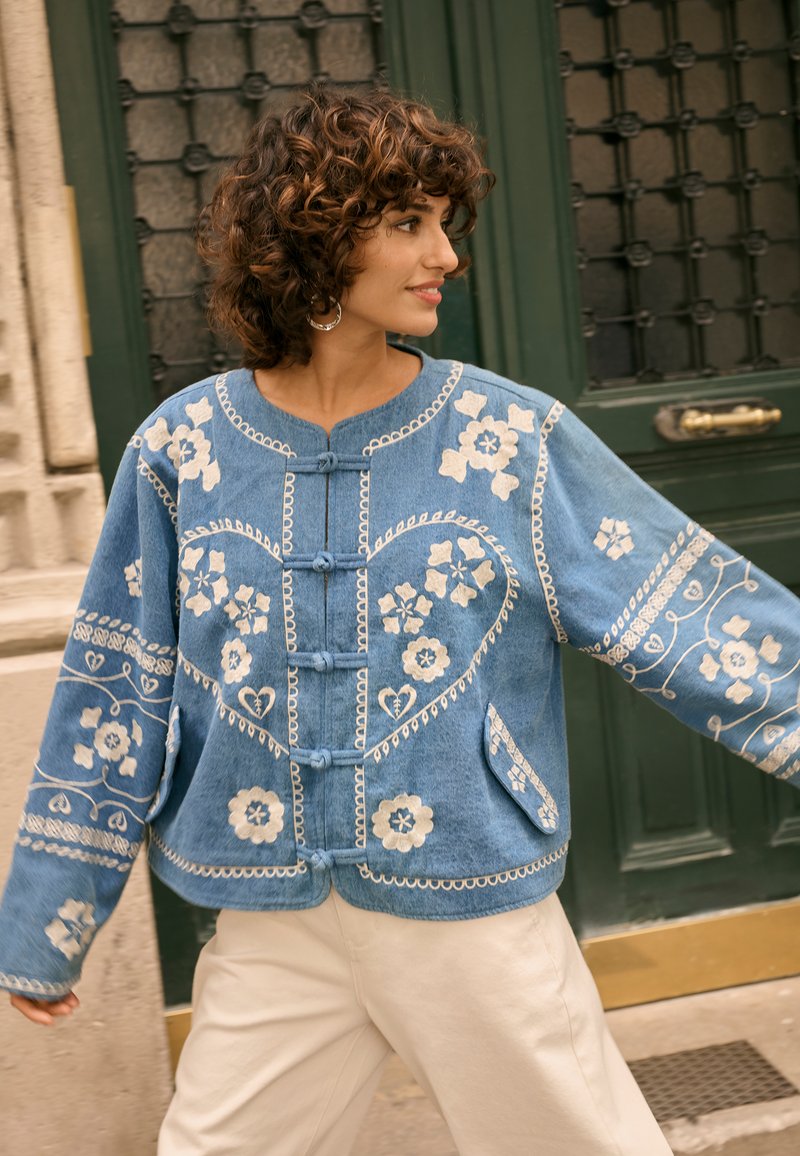 Ženska s kodrastimi lasmi, oblečena v modro jakno z belo cvetlično vezenino, stoji pred temno zelenimi vrati in gleda postrani.