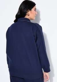 Donna con capelli scuri che indossa una maglia a maniche lunghe blu navy con dettagli cuciture bianche visibili, vista da dietro e di profilo.