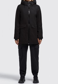 Sort hættetrøje parka med lynlås, to frontlommer og en glat, mat finish. Båret med matchende cargo bukser og mørkt fodtøj.