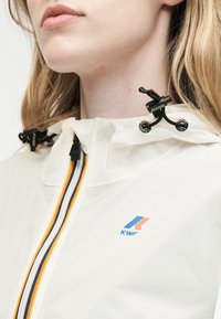 Κοντινό πλάνο ενός ατόμου που φοράει λευκό μπουφάν K-Way με ριγωτό φερμουάρ και ρυθμιζόμενα μαύρα κορδόνια κουκούλας.