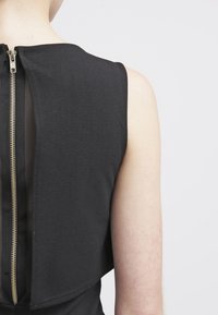 Vestido negro con una cremallera trasera prominente con accesorios dorados, material estructurado y un diseño en capas que muestra un panel transparente debajo.