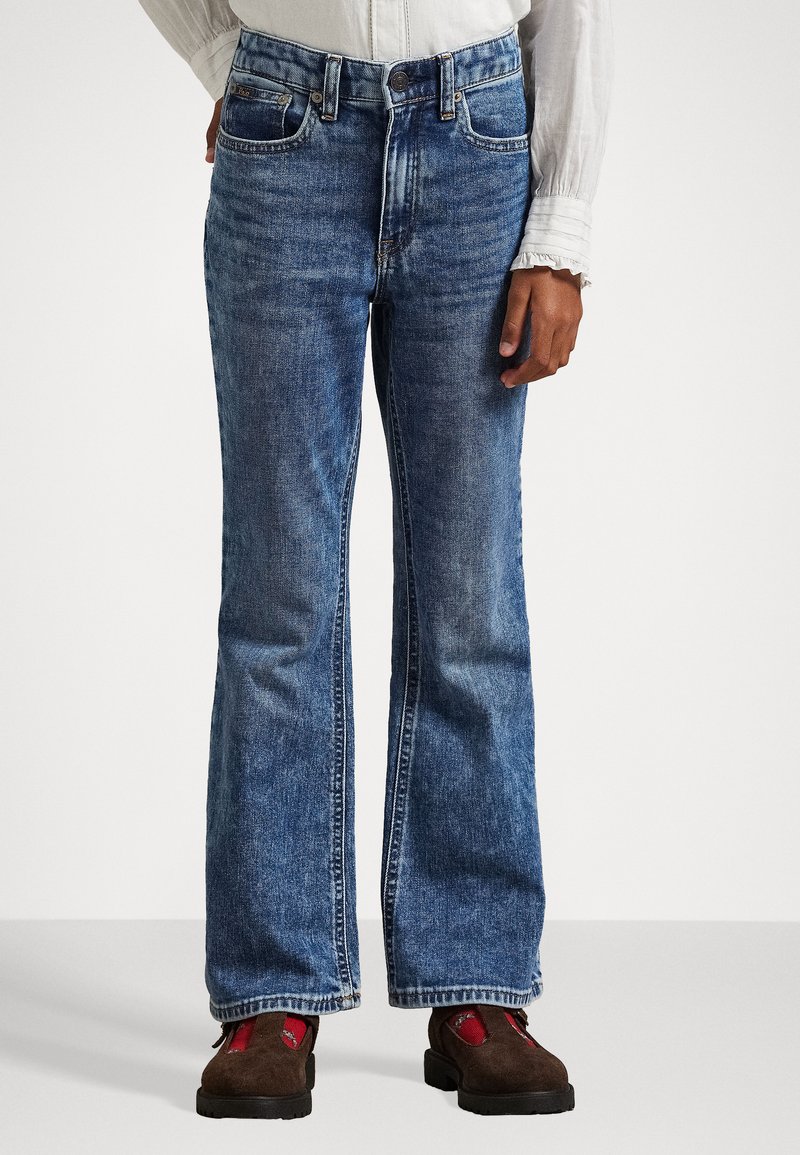 Blue denim flared jeans feature a classic cut, five pockets, and a button closure. The fabric has a slightly worn texture.