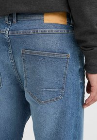 Jeans din denim albastru cu croială dreaptă, cu un buzunar tip patch și detalii subtile de cusătură. Talie are o etichetă din piele maro.