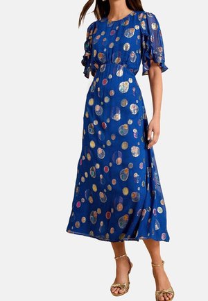 Kobieta ubrana w niebieską sukienkę midi z wielokolorowymi metalicznymi groszkami, bufkami na rękawach i złote sandały na wysokim obcasie z paskami.