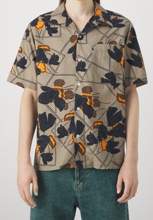 Chemise beige à manches courtes avec boutons, ornée d'un motif floral noir et orange, portée avec un jean bleu.