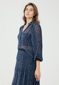 Marineblouse van doorschijnende stof, met pofmouwen en een kraag. Heeft een stippenpatroon in groen en paars. Gecombineerd met een geplisseerde rok.