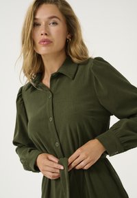 Robe chemise côtelée vert olive avec un col, fermeture frontale à boutons, manches bouffantes et lien à nouer à la taille, fabriquée dans un tissu texturé.