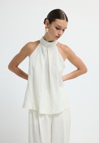 Haut à col halter blanc avec un col haut et un devant plissé. Tissu lisse avec une coupe ample, associé à un pantalon large assorti.