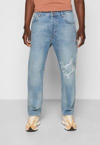 Ljusblå denimjeans med rak passform, med vit text "Pegador" på vänster ben. Bärs med krämfärgade och orange sneakers.