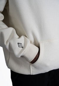 Hoodie color crema realizzato in un tessuto morbido, con polsini a coste e un logo geometrico ricamato sulla manica.