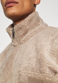 Beige fleece jakke med høj krave, halv lynlås og tekstureret overflade. Synlige sømme tilføjer detaljer til designet.