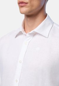 Camicia bianca in lino con colletto a punta, chiusura frontale con bottoni e discreto dettaglio del logo; tessuto leggero e design classico.