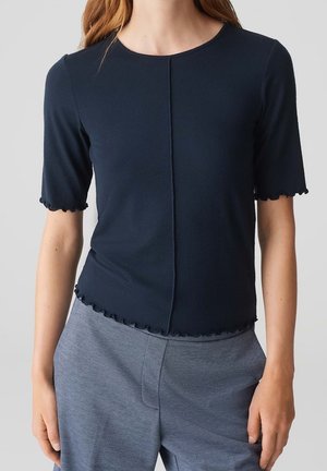 Femme portant un haut bleu marine ajusté à manches courtes avec des bords festonnés et un pantalon bleu ciel texturé.