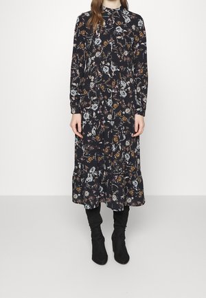 Femme portant une robe midi noire à manches longues avec un motif floral multicolore et des bottes à talons noirs, debout devant un fond blanc.