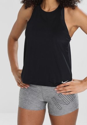 Femme portant un haut de sport noir sans manches et un short gris Nike, mains sur les hanches devant un fond uni.